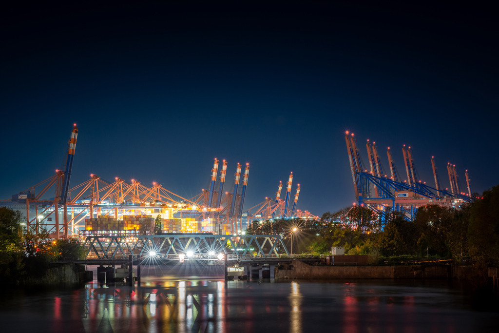 Roßkanal mit Blick auf die Container Terminals | Das Bild zeigt eine nächtliche Szenerie im Hamburger Hafen am Burchardkai, wo mehrere riesige Containerschiffe angedockt sind. Die Schiffe erheben sich majestätisch in die Dunkelheit und werden von einer Flut von Lichtern beleuchtet, die vom Hafen und von den Schiffen selbst ausgehen. Das Bild fängt die Hektik und den Puls des Hafens in der Nacht ein und vermittelt einen Eindruck von der enormen Größe und dem Einfluss dieses wichtigen Handelszentrums.