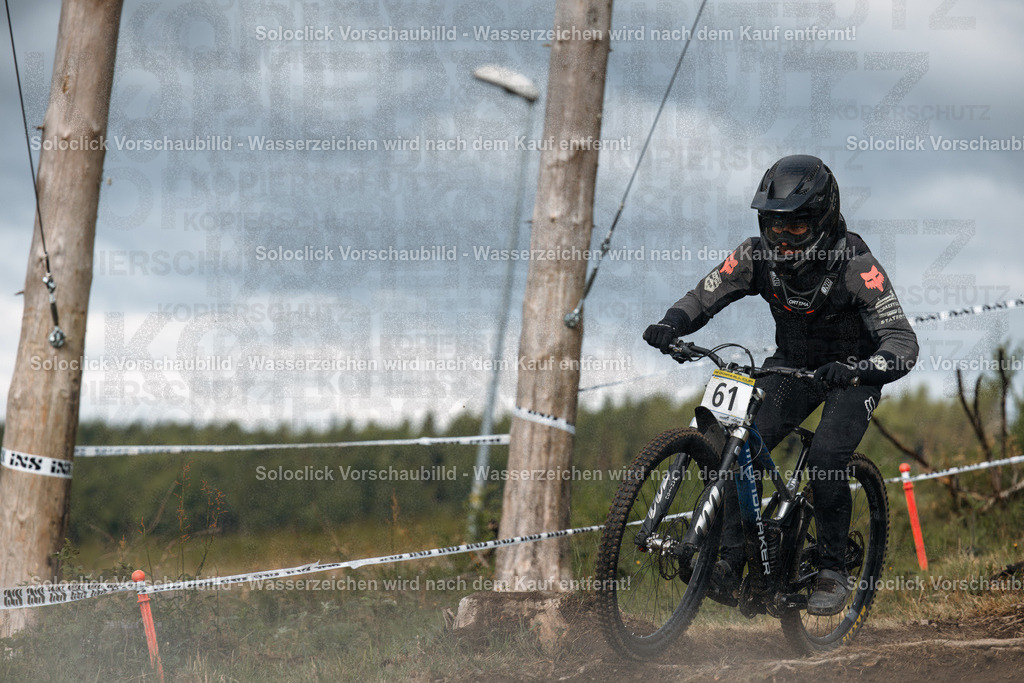 Action im Bikepark: Downhill-Rennen im Winterberg | Der Fotograf hat einen Mountainbiker in voller Aktion eingefangen, der einen staubigen Downhill-Kurs durch den Bikepark Winterberg durchquert. Der Fahrer trägt einen schwarzen Helm und eine schwarze Schutzkleidung und fährt ein Mountainbike mit breiten Reifen und einer speziellen Federung. Der Kurs ist mit roten und weißen Markierungspylonen gesäumt, und der Hintergrund besteht aus Bäumen und Büschen. Der Himmel ist bewölkt, was auf eine kühle, feuchte Atmosphäre hindeutet. - Realisiert mit Pictrs.com