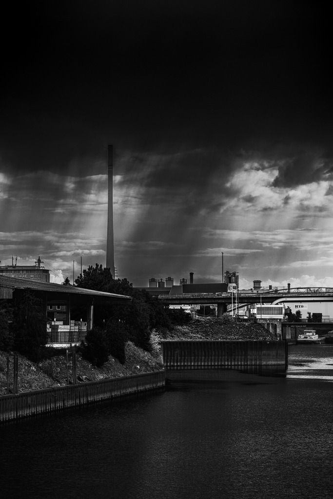 Zwischen Stahl und Sturm – Duisburger Hafen in Schwarzweiß | Dramatische Schwarzweißaufnahme aus dem industriellen Herzen des Ruhrgebiets. Dunkle Gewitterwolken ziehen über den Duisburger Hafen, während Schornsteine, Wasserwege und Industrieanlagen eine intensive urbane Atmosphäre erzeugen. Das Spiel aus Licht, Schatten und rauer Architektur verleiht dem Bild eine kraftvolle, fast filmische Stimmung – typisch Ruhrgebiet.Ideal als Fine-Art-Print für Liebhaber von Urban Photography, Industrieästhetik und authentischer Ruhrpott-Kultur. - Realisiert mit Pictrs.com