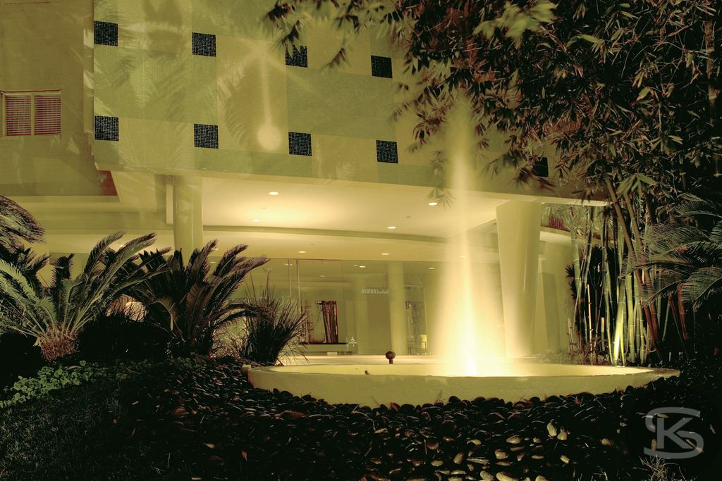 Nächtlicher Eingang des Avalon Hotel Beverly Hills mit Fontäne (Langzeitbelichtung, 2005) | Die nächtliche Außenaufnahme zeigt den hell beleuchteten, modernen Eingangsbereich des Avalon Hotel Beverly Hills, aufgenommen im Jahr 2005. Das Bild verwendet eine Langzeitbelichtung, um die sanfte Bewegung des Wassers in einer kreisförmigen Hotellobby-Fontäne einzufangen, umgeben von dunklen Büschen und Palmen, die einen Hauch von kalifornischem Chic und 1950er-Jahre-Flair ausstrahlen. - Realisiert mit Pictrs.com
