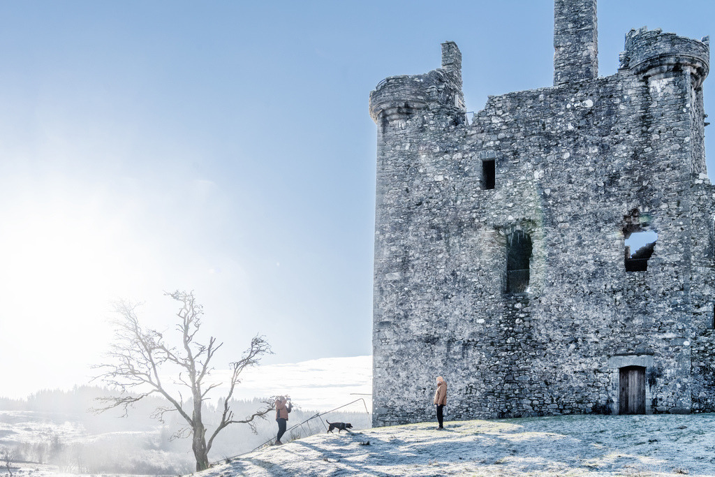 Kilchurn Castle Winterlandschaft Schottland | Die Ruine von Kilchurn Castle in Schottland präsentiert sich in einer frostigen Winterlandschaft. Zwei Personen und ein Hund erkunden die Umgebung des historischen Bauwerks. Das klare Mittagslicht betont die Textur der alten Steinmauern und die winterliche Atmosphäre. - Realisiert mit Pictrs.com