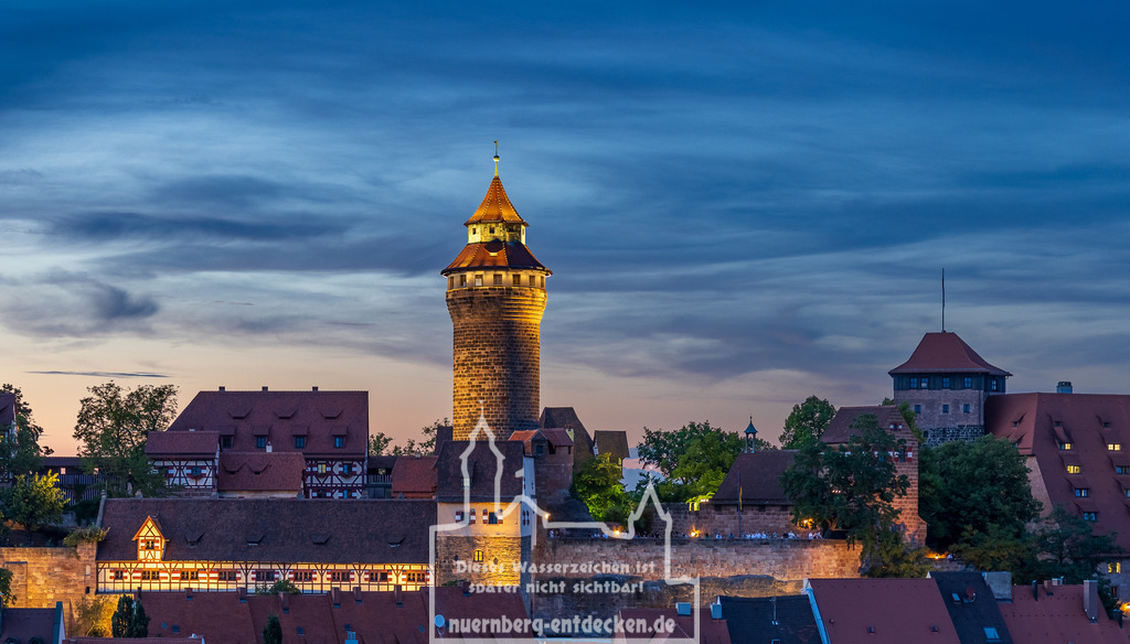 Monatsbild des Kalender 2025 für den Monat August | Die Kaiserburg in Nuernberg bei Dämmerung mit dem beleuchteten Sinwellturm im Zentrum, einem der Wahrzeichen der Stadt. Die historischen Gebaeude und die mächtigen Burgmauern erstrahlen im warmen Licht, waehrend der Himmel in Blau- und Orangetoenen den Uebergang von Tag zu Nacht andeutet. - Realisiert mit Pictrs.com