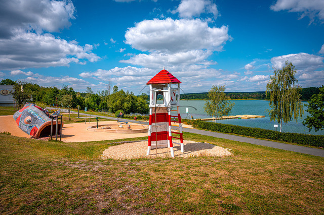 2026-07-28_11-56-06_810 | Leuchtturm auf Spielplatz am Seeufer Freibad Zeulenrodaer-Meer unter blauem Himmel mit Wolken. Sommerliche Atmosphäre und grüne Umgebung, Zeulenroda-Triebes, Thüringen, Deutschland - Realisiert mit Pictrs.com