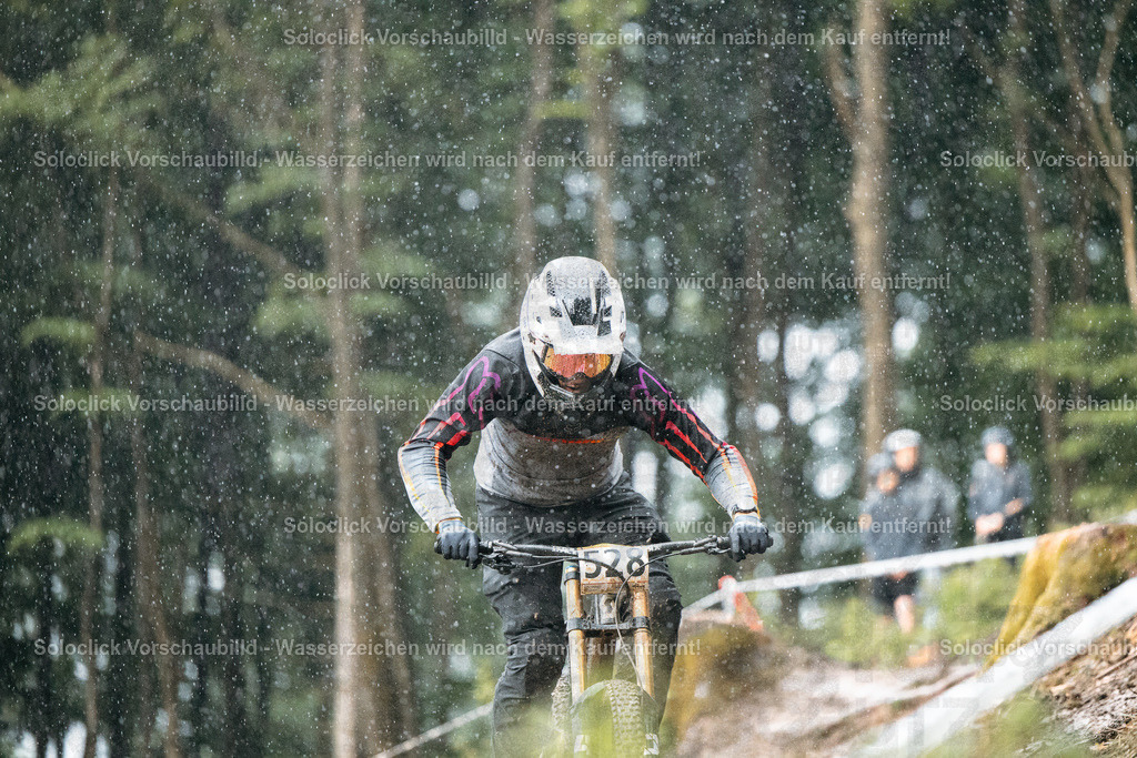 Adrenalin im Wald | Ein Mountainbiker rast in voller Geschwindigkeit einen staubigen, schmalen Pfad in einem dichten Wald hinunter. Der Fahrer trägt einen weißen Helm und eine dunkle Schutzkleidung. Der Pfad ist mit Wurzeln und Steinen übersät, was die Fahrt anspruchsvoll macht. Im Hintergrund sind weitere Bäume und ein paar Zuschauer zu sehen. - Realisiert mit Pictrs.com
