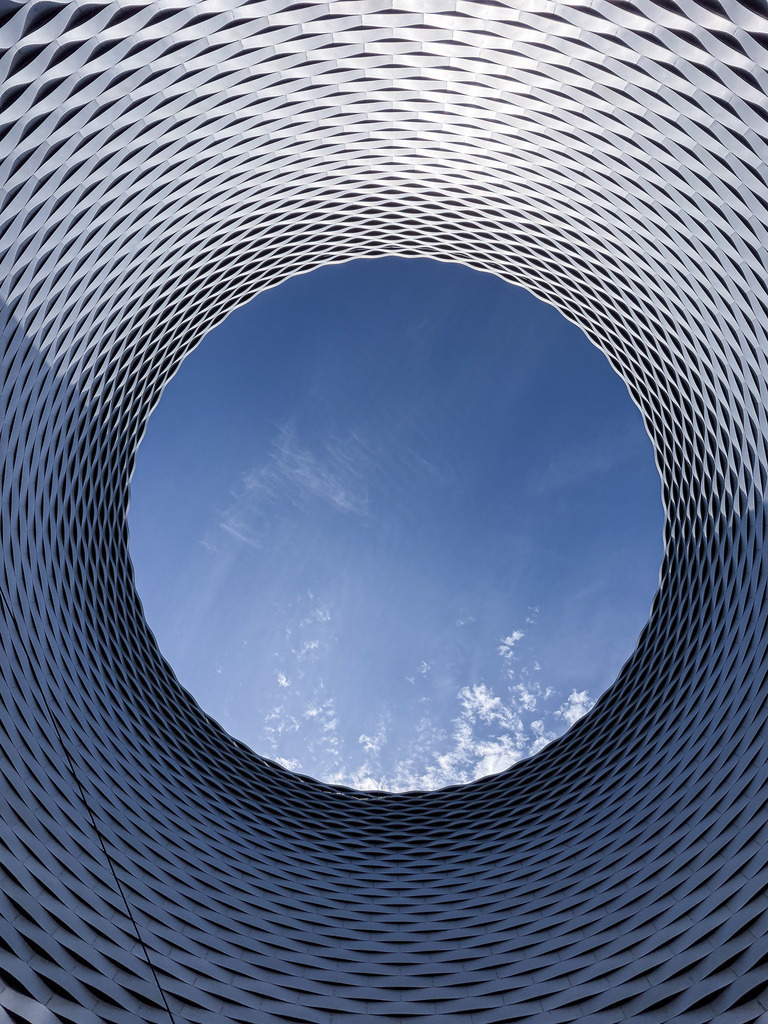 Wandbild Messe Basel Himmelsloch – Moderne Architekturfotografie | Ein meisterhafter Blick durch das Herz der modernen Architektur: Diese perfekt zentrierte Fine-Art-Aufnahme fängt die weltberühmte, geschwungene Aluminium-Fassade der Messe Basel (Herzog &amp; de Meuron) ein. Der spektakuläre Kontrast zwischen der kühlen, mathematischen Metallstruktur und dem lebendigen, blauen Himmel mit sanften Wolken macht dieses Werk zu einem ultimativen Blickfang. Perfekt für moderne Wohnräume, Lofts oder Architektur-Büros. Empfohlen als Großformat unter Acrylglas für maximale Tiefenwirkung. Jetzt als hochwertiges Poster oder Premium-Kunstdruck für deine Wandgestaltung sichern! - Realisiert mit Pictrs.com