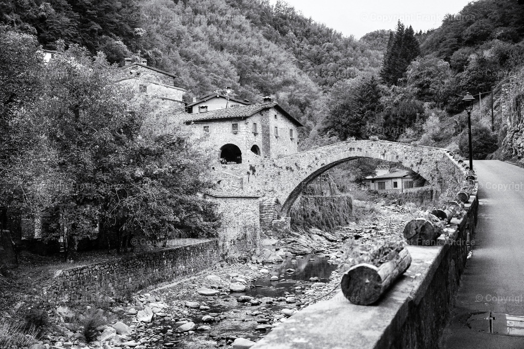 Alte Steinbrücke in der Toskana | Schwarz weiss Fotografie von der Ponte Della Dogana, eine alte Steinbrücke in Fabbriche di Vergemoli Toskana. Die Brücke führt über einen kleinen Fluss zu alten Häusern. Im Hintergrund die Berge der Toskana in der Region Lucca. - Realisiert mit Pictrs.com