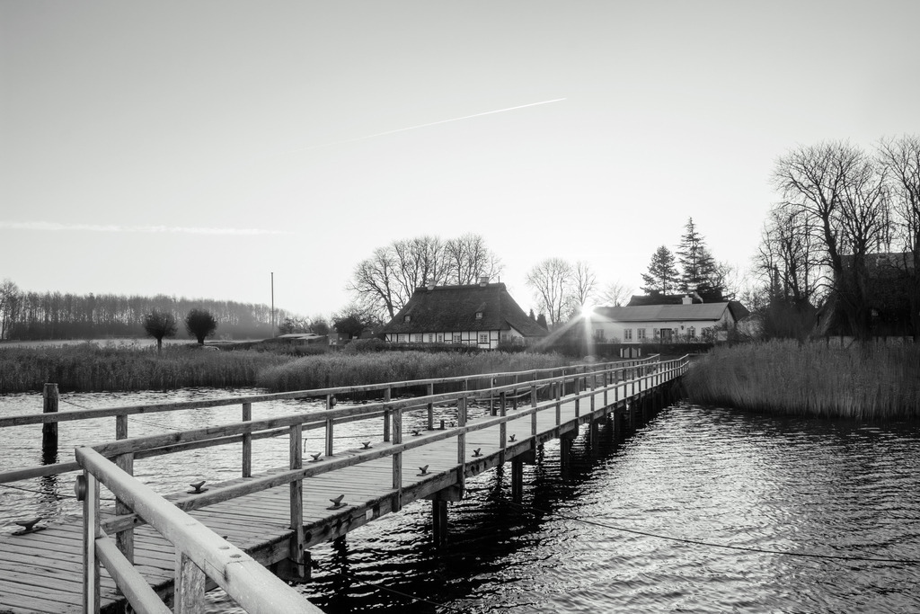 Wandbild: Sonnenaufgang in Sieseby in Schwarz-Weiß | Dieses Wandbild im Querformat zeigt einen schönen Sonnenaufgang in Sieseby in Schwarz-Weiß. Im Vordergrund ist der Schleianleger zu sehen. Die Sonne geht zwischen den beiden malerischen Häusern am Schleiufer auf. Auch die Sonnenstrahlen sind gut zu erkennen. Am Horizont ist der Himmel durch die morgendliche Lichtstimmung schön hell. Der Himmel ist fast wolkenlos.  - Realisiert mit Pictrs.com