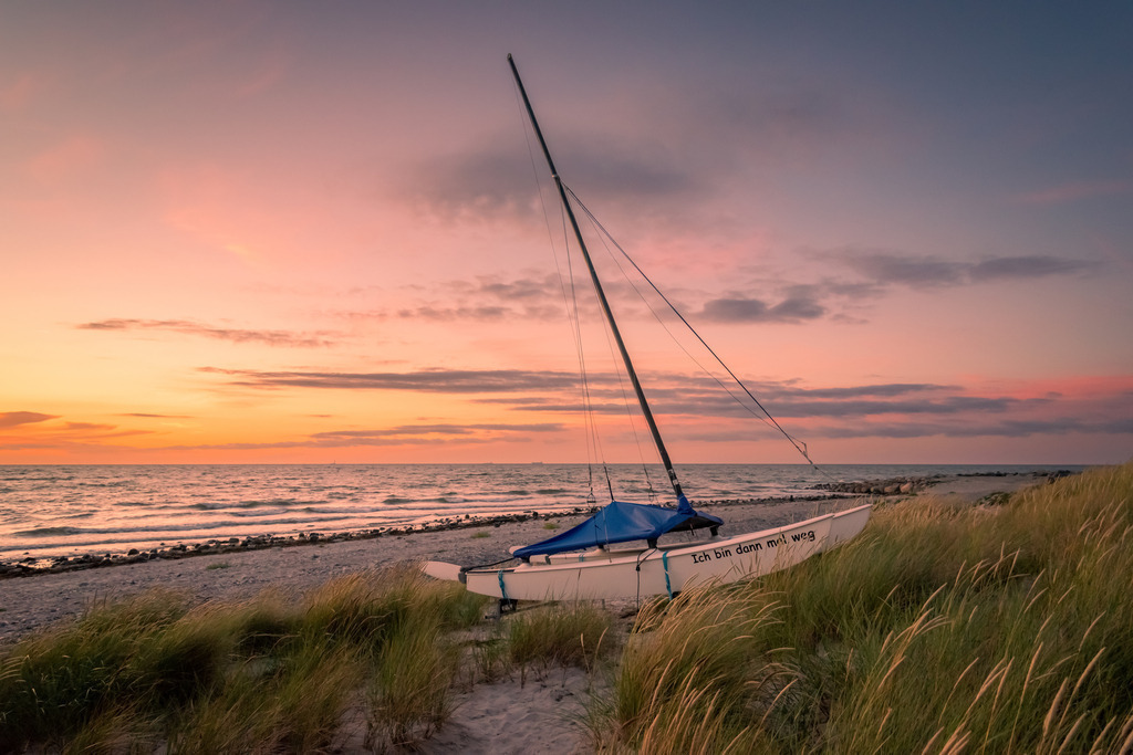 Katamaran am Strand von Bottsand | Die Sonne, die gerade dabei ist unterzugehen, taucht die Szene in ein warmes Licht, das dem ganzen Bild eine romantische und friedliche Atmosphäre verleiht. Die Szenerie ist vollkommen still und friedlich, und man kann förmlich das Rauschen des Meeres hören.