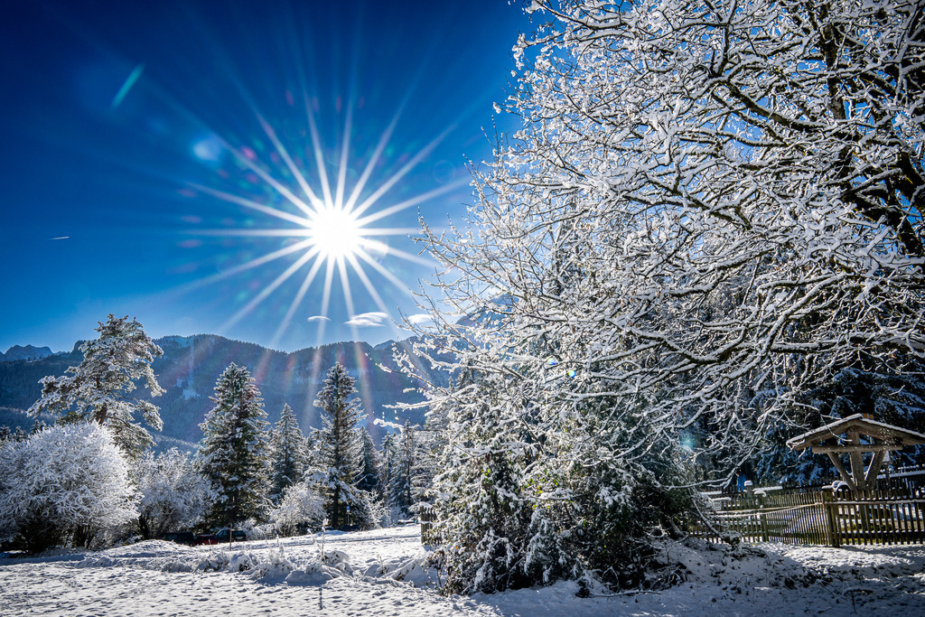 Winterzauber in Garmisch-Partenkirchen: Sonnenstern über tiefverschneiter Bergwelt | Ein strahlender Sonnenstern lässt die winterliche Alpenlandschaft rund um Garmisch-Partenkirchen in magischem Glanz erstrahlen. Unter einem makellos blauen Himmel glitzert der frisch gefallene Schnee auf den verschneiten Baumkronen und der weiten, unberührten Fläche im Vordergrund. Das Zusammenspiel aus eisiger Frühlings- beziehungsweise Winterluft, glitzerndem Weiß und der wärmenden Intensität der Sonne erzeugt eine friedliche, fast märchenhafte Atmosphäre, die pure Entschleunigung und Winterromantik ausstrahlt. - Realisiert mit Pictrs.com