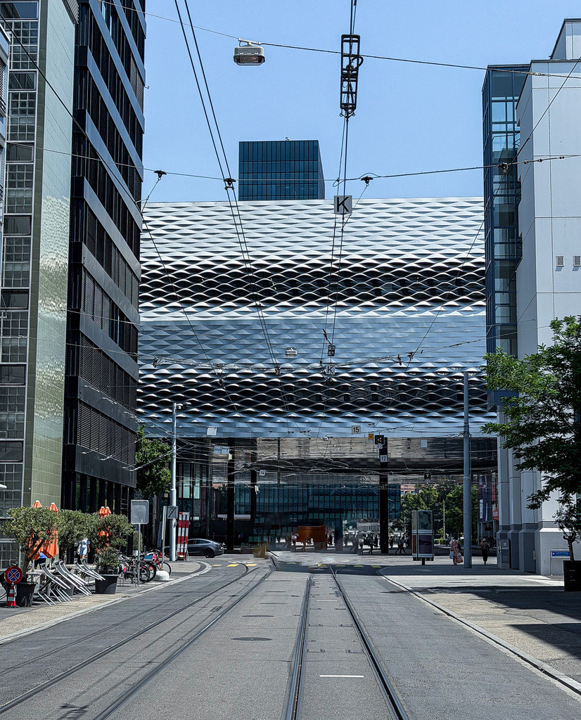 Wandbild Moderne Fassadenkunst Basel - Urbane Architektur-Fotografie | Dieses dynamische Architektur-Wandbild fängt den modernen, urbanen Puls der Stadt Basel ein. Die beeindruckende, spiegelnde Glasfassade und das geometrische Linienspiel im Zusammenspiel mit den Fluchtlinien der Straße und Oberleitungen erzeugen eine faszinierende räumliche Tiefe. Fotograf Robert Steinke inszeniert diese kosmopolitische Szene mit einem präzisen Blick für Symmetrie und zeitgenössisches Urban-Design. Ein absolut ausdrucksstarker Blickfang für moderne Wohnzimmer, Loft-Wohnungen oder repräsentative Büroräume, die eine weite, architektonische Eleganz ausstrahlen sollen. Perfekt geeignet als großformatiger Premium-Kunstdruck auf edlem Alu-Dibond, Leinwand oder unter brillantem Acrylglas für maximale Tiefenwirkung. - Realisiert mit Pictrs.com