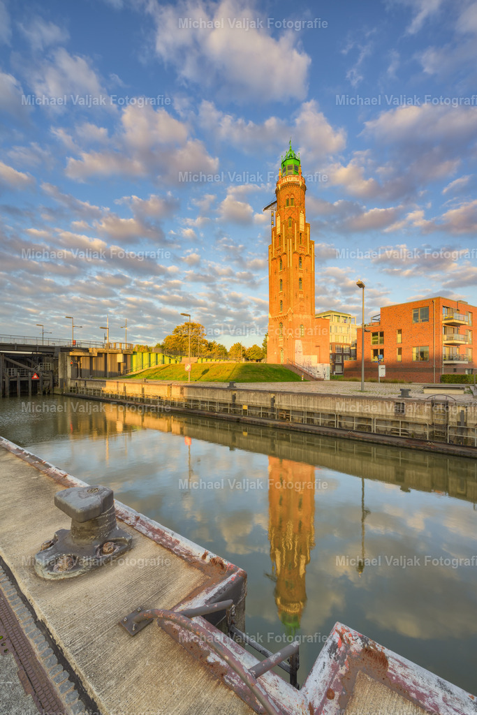 Leuchtturm Bremerhaven im Morgenlicht | Ein stiller Moment am Neuen Hafen in Bremerhaven: Der Simon-Loschen-Leuchtturm, ältester Festland-Leuchtturm an der deutschen Nordseeküste, wird von der ersten Morgensonne in warmes Licht getaucht. Die neugotischen Formen des Backsteinbaus leuchten rötlich auf, während sich sein schlanker Turm im ruhigen Wasser der Schleuse spiegelt. - Realisiert mit Pictrs.com