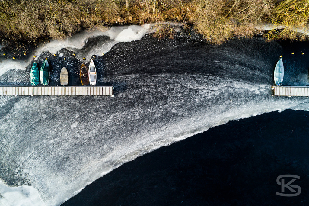 Eingefrorene Boote am Wintersteg – Drohnenaufnahme eines zugefrorenen Sees | Beeindruckende Luftaufnahme von eingefrorenen Booten am Steg eines zugefrorenen Sees. Die Drohnenfotografie zeigt winterliche Stille mit Baumschatten auf der Eisfläche aus der Vogelperspektive – atmosphärische Winterimpression. - Realisiert mit Pictrs.com