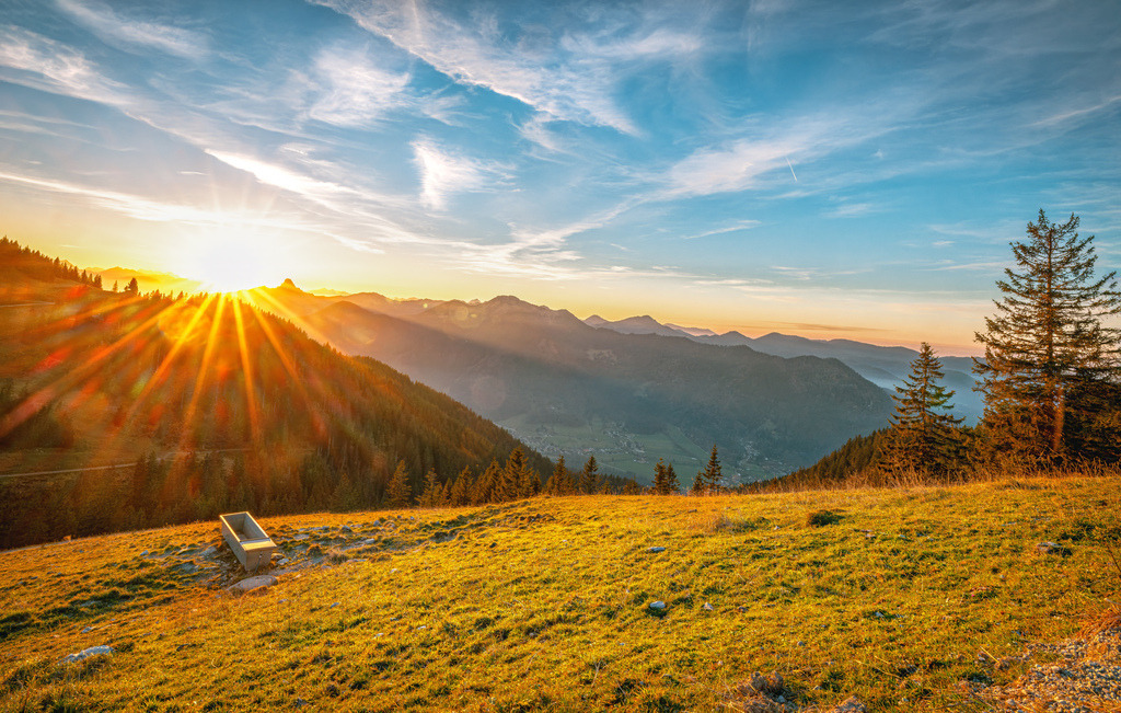 Herbstlicher Sonnenuntergang über die Oberbayrische Berglandschaft des Tegernsee's | Das Bild zeigt einen malerischen Sonnenuntergang in einer herbstlichen Berglandschaft. Es handelt sich um eine Aufnahme, die wahrscheinlich im bayerischen Alpenraum, möglicherweise in der Nähe des Tegernsees, entstanden ist. Die Szenerie zeigt verschneite Hügel im Vordergrund und eine Bergkette im Hintergrund, beleuchtet durch die tief stehende Sonne.Die Aufnahme fängt dramatische Wolkenformationen und intensive Lichtstrahlen ein. - Realisiert mit Pictrs.com
