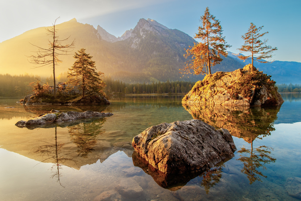 Morgenruhe am Hintersee - Farben der Stille | Dieses Wandbild zeigt die beeindruckende Schönheit des Hintersees in Bayern im ersten Licht des Tages. Die goldene Morgensonne taucht die kleinen Felseninseln mit ihren Bäumen in warmes Licht. Die spiegelglatte Wasseroberfläche reflektiert Felsen, Bäume und Berge in perfekter Klarheit. Eine Szene voller Frieden und natürlicher Harmonie. Ideal für Wohnräume, Praxis, Büro oder Schlafzimmer. Dieses Motiv strahlt Ruhe, Weite und Erdung aus und bringt die Kraft der Natur direkt in dein Zuhause.