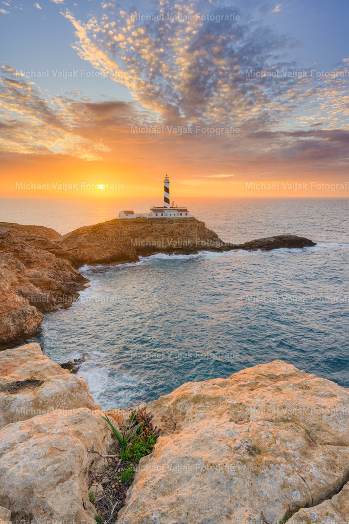 Sonnenaufgang am Faro de Cala Figuera auf Mallorca | Im Vordergrund liegen die hellen, rauen Felsen, dahinter öffnet sich die geschützte Bucht mit ihrem ruhigen Wasser, bevor der Blick weiter zum Faro de Cala Figuera und dem aufsteigenden Sonnenlicht führt. Der Himmel leuchtet in satten Orange‑ und Gelbtönen, die sich klar über der Küstenlinie abzeichnen und sowohl die Felsen als auch den Leuchtturm in ein gleichmäßiges, warmes Morgenlicht setzen. Die vertikale Perspektive betont die Abfolge von Fels, Wasser und Küste und zeigt den Sonnenaufgang als klar strukturierten, sachlichen Moment. - Realisiert mit Pictrs.com