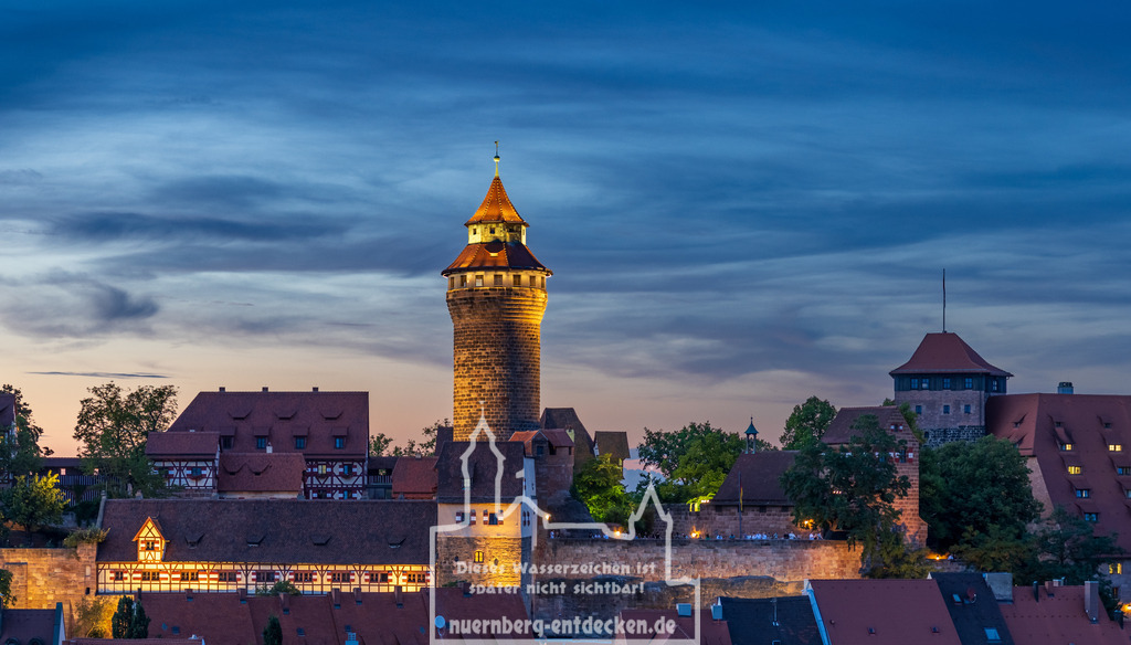 Nürnberger Stadtbild am Abend | Die Kaiserburg in Nürnberg bei Dämmerung mit dem beleuchteten Sinwellturm im Zentrum, einem der Wahrzeichen der Stadt. Die historischen Gebäude und die mächtigen Burgmauern erstrahlen im warmen Licht, während der Himmel in Blau- und Orangetönen den Übergang von Tag zu Nacht andeutet. - Realisiert mit Pictrs.com