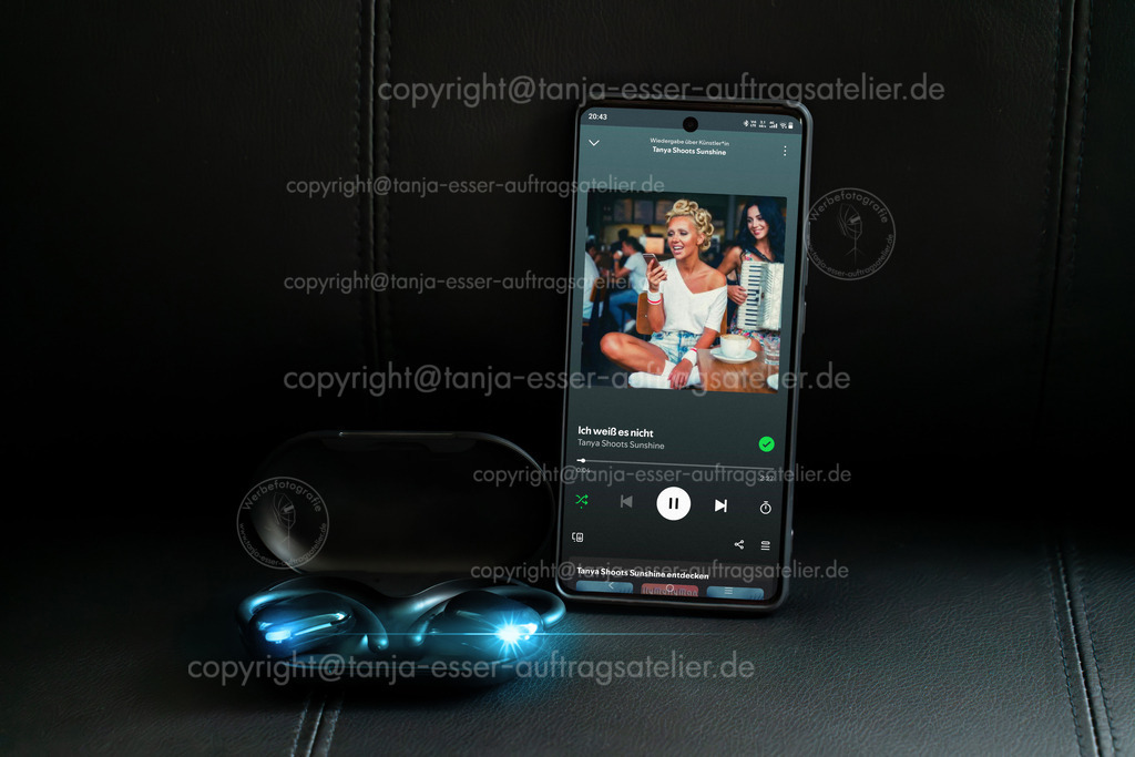 Kopfhörer mit Smartphone  zeigt einen Song Editorial | Brilon, Nordrhein-Westfalen, Deutschland – 15. Oktober 2026: Ein Smartphone zeigt Spotify App mit dem Song „Ich weiß es nicht“ von Tanya Shoots Sunshine. Daneben liegen moderne Open-Ear Bluetooth-Kopfhörer in einem Ladeetui auf dunklem Ledersessel.