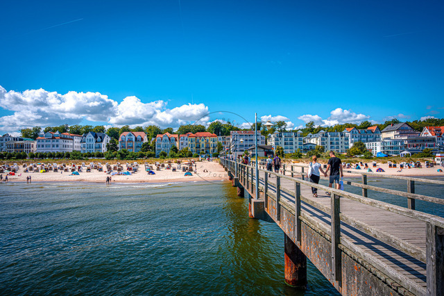 2026-08-08_11-53-04_840 | Ein langer Pier führt zum Strand mit bunten Häusern im Hintergrund unter einem blauen, wolkenbedeckten Himmel, Bansin, Usedom, Mecklenburg-Vorpommern, Deutschland - Realisiert mit Pictrs.com