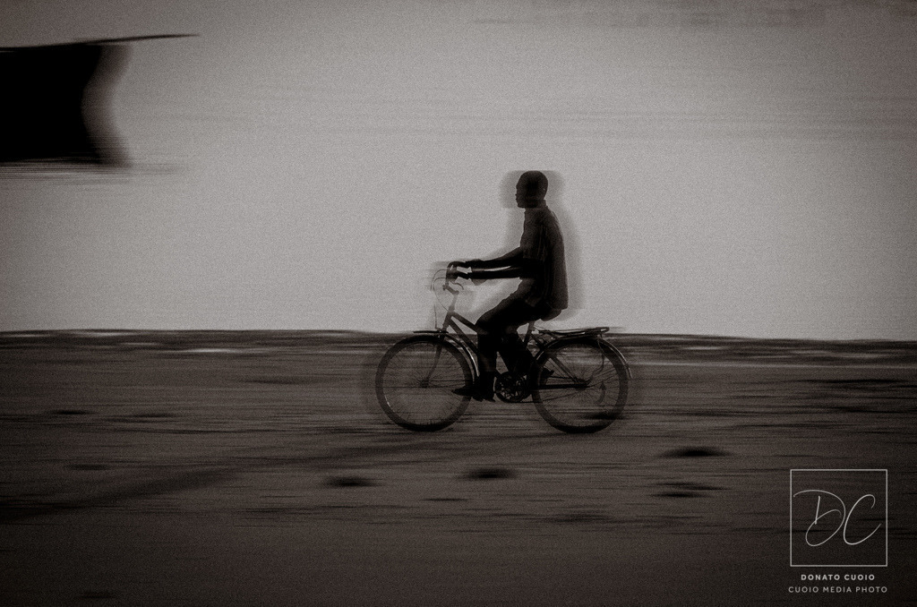 Der Weg zur Arbeit – Sansibar im Morgengrauen | Dieses eindrucksvolle Schwarz-Weiß-Fotografie wurde am frühen Morgen am Strand von Sansibar aufgenommen. Es zeigt eine Person auf dem Fahrrad, die auf dem Weg zur Arbeit ist und die Weite des Strandes durchquert. Durch die nachträgliche digitale Bearbeitung mit Bewegungsunschärfe entsteht eine besondere Dynamik, die das Gefühl von Geschwindigkeit und Vergänglichkeit verstärkt. Die monochrome Ästhetik verleiht dem Bild eine zeitlose Eleganz und unterstreicht die Atmosphäre des Augenblicks.Ein einzigartiges Kunstwerk für Liebhaber von Street Photography, Reisefotografie und minimalistischer Ästhetik. Perfekt als Wandbild, Fine-Art-Print oder stilvolle Dekoration für moderne Wohnräume. - Realisiert mit Pictrs.com