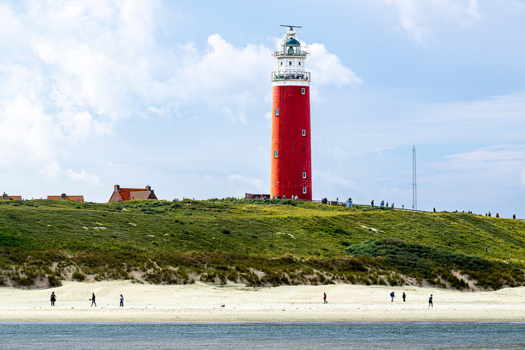 Wahrzeichen der Nordseeinsel Texel: Roter Leuchtturm Eierland über den Dünen bei De Cocksdorp | Der leuchtend rote Leuchtturm Eierland ragt majestätisch hinter begründen Dünen an der Nordspitze der niederländischen Insel Texel bei De Cocksdorp empor. Auf dem Weg um den Turm sowie am feinsandigen Strand im Vordergrund sind Spaziergänger unterwegs. Ein leicht bewölkter Sommerhimmel komplettiert die klassische Küstenszenerie. Ideal nutzbar für redaktionelle Beiträge zu Themen wie Nordseetourismus, Wahrzeichen der Niederlande, Inselurlaub und Küstenarchitektur. - Realisiert mit Pictrs.com