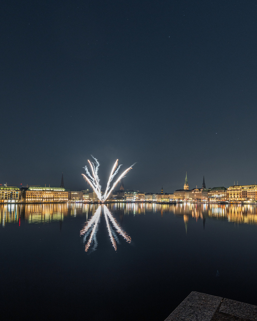 Feuerwerk über der Hamburger Alster | Die ersten Feuerwerksfontänen steigen über der nächtlichen Hamburger Alster auf und spiegeln sich im ruhigen Wasser. Die beleuchtete Innenstadt bildet eine elegante Kulisse. - Realisiert mit Pictrs.com