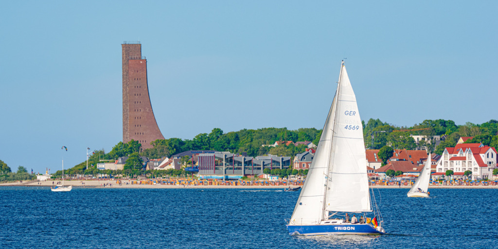 Laboe - Kieler Förde | Segelboote im Sommer auf der Kieler Förde; im Hintergrund präsentieren sich der weite Strand, ansehnliche Häuser und das Marine-Ehrenmal in Laboe. — Auflösung des Originals: 6016 x 3008 px. - Realisiert mit Pictrs.com