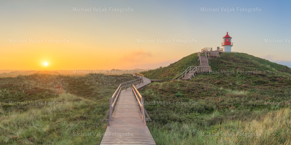 Morgens beim Quermarkenfeuer Norddorf auf Amrum | Ein weiter Morgenhimmel spannt sich über die Dünen von Amrum. Links steigt die Sonne langsam empor und taucht die Landschaft in sanftes Gold. Der Holzsteg in der Bildmitte zieht sich ruhig durch die Heide – eine Linie der Orientierung in der Weite. Rechts, auf erhöhter Düne, steht das Quermarkenfeuer: schlicht, funktional, und doch markant. Das Panorama verbindet Licht, Weg und Landmarke zu einem stillen Bild maritimer Klarheit. - Realisiert mit Pictrs.com