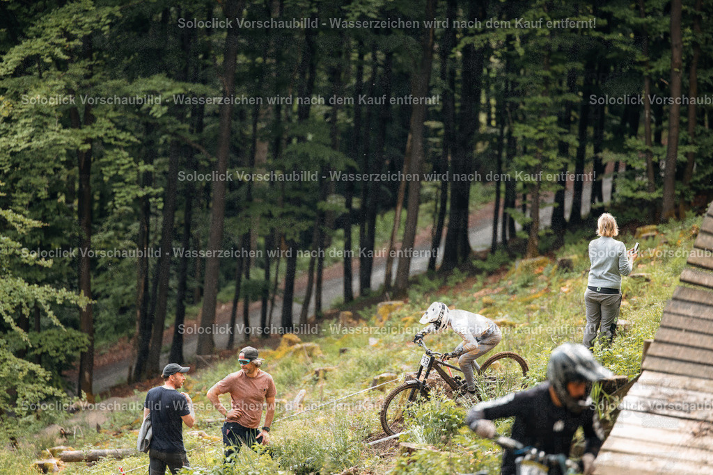Mountainbike-Action im Wald | Die Aufnahme zeigt eine Szene im Herzen eines dichten, grünen Waldes. Mehrere Personen, vermutlich Mountainbiker und Betreuer, sind in der Szene zu sehen. Einige Biker stehen mit ihren Fahrrädern, während andere sich beraten oder die Strecke inspizieren. Die Atmosphäre ist konzentriert und sportlich. - Realisiert mit Pictrs.com