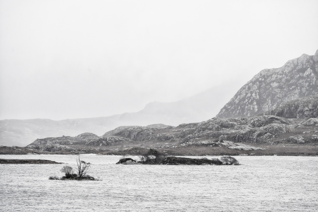 Loch Tollaidh | Eine weite Wasserfläche liegt unter einem hellen, wolkenverhangenen Himmel. Im Vordergrund befinden sich zwei kleine, felsige Inseln, die spärlich mit blattlosen Bäumen und Sträuchern bewachsen sind. Das Wasser kräuselt sich leicht im Wind. Im Hintergrund erheben sich die schroffen, dunklen Bergketten von Wester Ross. Die Gipfel sind teilweise von tief hängenden Wolken und Dunst verschleiert, was der kargen Landschaft eine ruhige, melancholische Atmosphäre verleiht. Die fast monochrome Aufnahme betont die harten Kontraste der Felsformationen und die einsame Weite der schottischen Natur. - Realisiert mit Pictrs.com