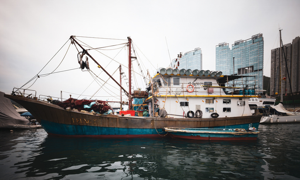 Ein traditionelles Fischerboot liegt ruhig im Hafen von Hongkong | Das Bild zeigt ein traditionelles Fischerboot mit einem weißen Aufbau und einer Reihe von runden Lichtern oder Netzen an Bord, das an einem Kai oder in einem Hafen von Honkong im Wasser liegt - Realisiert mit Pictrs.com