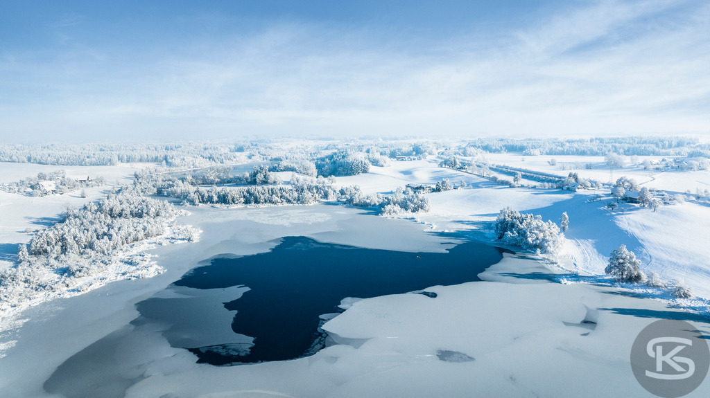 Gefrorener See im Winter – Eislandschaft aus der Vogelperspektive | Diese Drohnenaufnahme zeigt einen teilweise zugefrorenen See in verschneiter Winterlandschaft. Offene Wasserflächen bilden dunkle Kontraste zur weißen Eisfläche und den schneebedeckten Ufern. Die Luftperspektive offenbart die grafische Qualität der gefrorenen Strukturen und die klaren Linien zwischen Eis, Wasser und verschneitem Wald unter blauem Winterhimmel. - Realisiert mit Pictrs.com