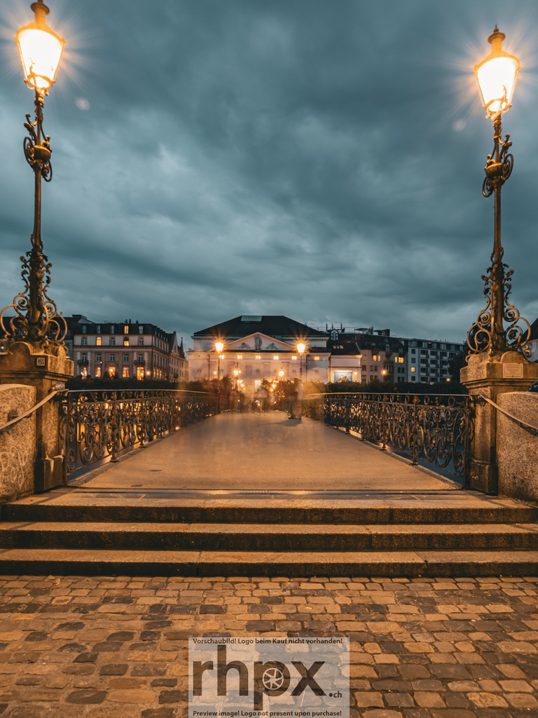 Abend am Rathaussteg | Licht. Symmetrie. Verbindung.
Der beleuchtete Rathaussteg führt den Blick direkt auf das Luzerner Theater unter einem dramatischen Abendhimmel. Die filigranen Verzierungen der Laternen rahmen diesen urbanen Moment perfekt ein und laden zum nächtlichen Flanieren ein.
Wähle unter "Produktauswahl" dein Wunschformat: Vom klassischen Wandbild über Puzzle &amp; Tassen bis zum digitalen Download (z.B. als Handy-Hintergrund).
 - Realisiert mit Pictrs.com
