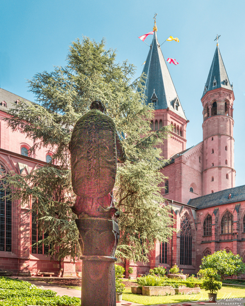 Der Kreuzgang des Mainzer Doms mit Sandsteinfigur | Die Sandsteinfigur eines in liturgische Gewänder gehüllten Heiligen mit Buch in der Hand. Die andere Hand hielt vermutlich ein spezielleres, identifizierendes Attribut, welches leider verloren ist. Daher ist die Identifizierung des Heiligen heute eher wage. Es könnte sich um einen hl. Stephanus handeln, allerdings fehlen die attributiven Steine. In jedem Fall ist die Figur in die Zeit des Spätbarock (18. Jh.) zu datieren und steht im Garten des Kreuzgangs des Mainzer Doms.
