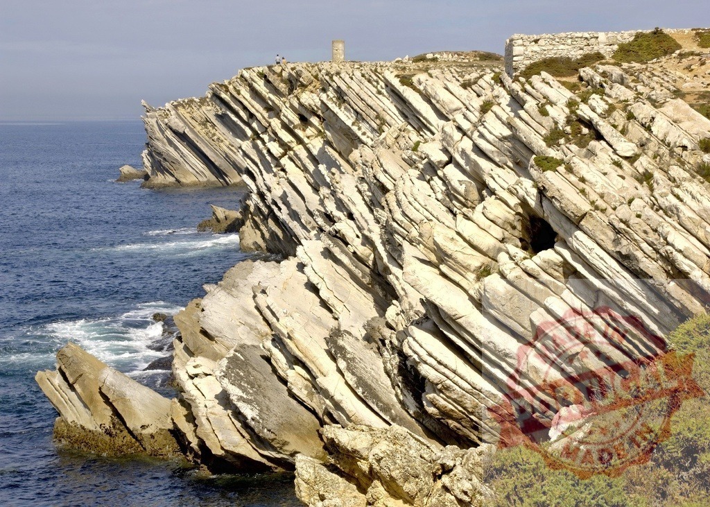 Layered sedimentary rock cliffs forming a rugged coastline at Peniche | Atlantic Ocean waves washing onto steep, layered cliffs with a historical tower in the distance - Realized with Pictrs.com