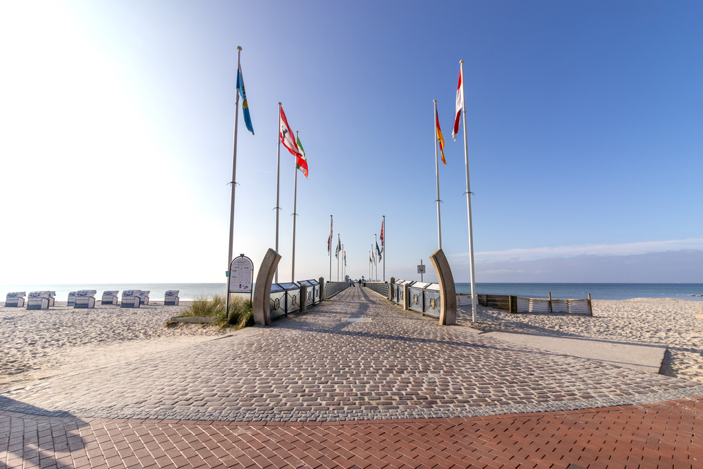 Wandbild: Promenade zur Seebrücke – Grömitz am Strand | Dieses Wandbild zeigt den gepflegten Strandzugang von Grömitz an einem sonnigen Tag. Eine breite Promenade aus roten Pflastersteinen führt geradlinig durch die Dünenlandschaft direkt auf die Seebrücke, die sich weit ins Meer erstreckt. Links und rechts säumen Fahnenmasten mit internationalen und regionalen Flaggen den Weg und verleihen der Szene ein weltoffenes Flair. Am Ende der Promenade sind Strandkörbe in ordentlichen Reihen aufgestellt, alle ausgerichtet zum Wasser hin. Der Himmel ist klar, mit nur wenigen Wolken am Horizont, und das Meer liegt ruhig und einladend vor dem Betrachter. Die Komposition wirkt strukturiert und zugleich weitläufig – ein typisches Bild für die Ostseeküste, das sowohl Ordnung als auch Entspannung vermittelt. Dieses Motiv eignet sich ideal als Wandbild für maritime Wohnkonzepte – ob als Leinwandbild, Acrylglasbild, Alu-Dibond FineArt Print oder als Akustikbild. Ein stilvoller Akzent für Wohnzimmer, Büro oder Ferienwohnung. - Realisiert mit Pictrs.com