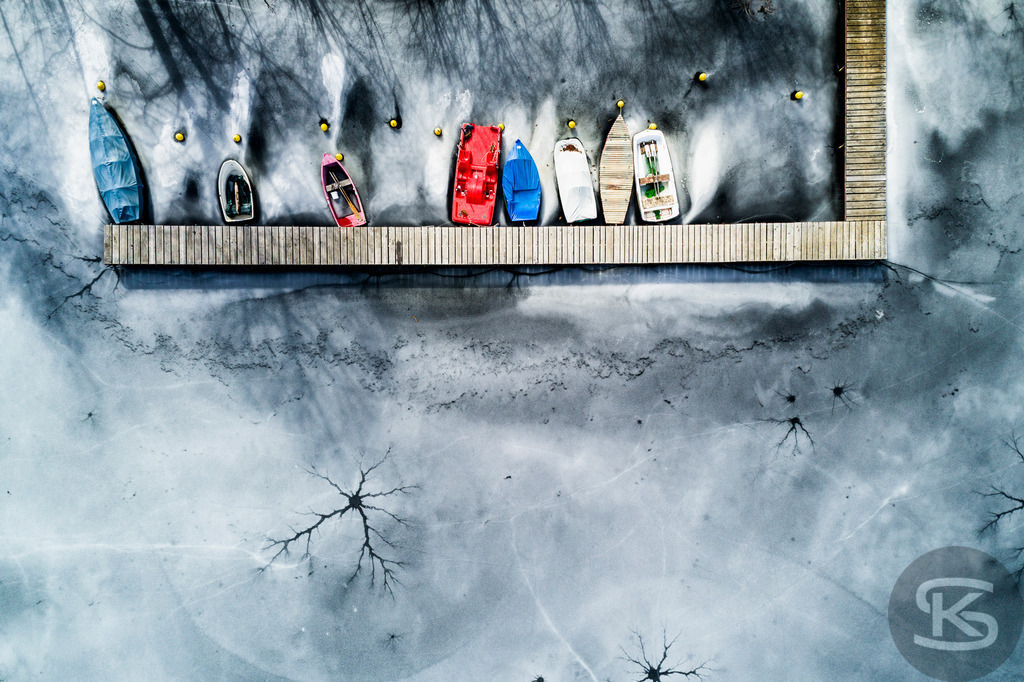 Eingefrorene Boote am Wintersteg – Drohnenaufnahme eines zugefrorenen Sees | Beeindruckende Luftaufnahme von eingefrorenen Booten am Steg eines zugefrorenen Sees. Die Drohnenfotografie zeigt winterliche Stille mit Baumschatten auf der Eisfläche aus der Vogelperspektive – atmosphärische Winterimpression. - Realisiert mit Pictrs.com