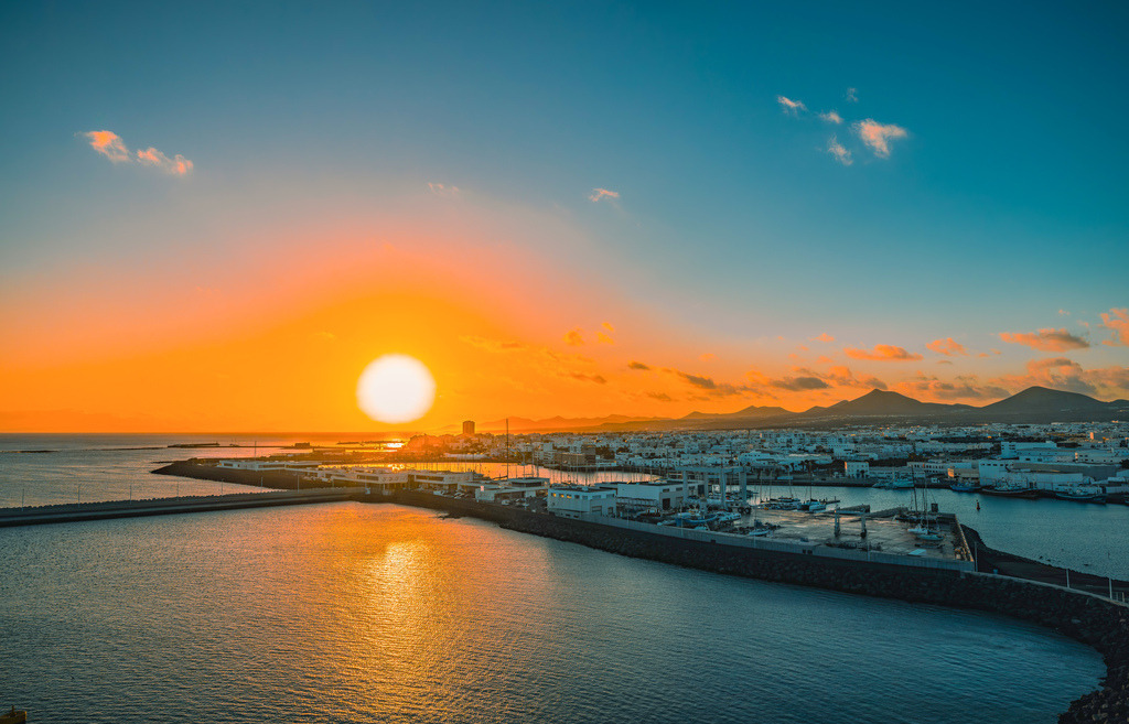 Sonnenuntergang über dem Hafen von Fuerteventura, sanfte Hügelketten am Horizont | Das Bild zeigt einen Sonnenuntergang über dem Hafen von Puerto del Rosario, der Hauptstadt der Kanareninsel Fuerteventura. Details zum AufnahmeortStandort: Der Blick geht über das Hafenbecken von Puerto del Rosario, wo häufig Kreuzfahrtschiffe anlegen.Merkmale: Im Vordergrund ist eine Hafenmole zu sehen, während sich im Hintergrund die Silhouette der kargen Vulkanlandschaft der Insel abzeichnet.Kontext: Puerto del Rosario ist das wirtschaftliche und administrative Zentrum Fuerteventuras und dient als wichtiger Knotenpunkt für den Schiffsverkehr zwischen den Inseln.  - Realized with Pictrs.com