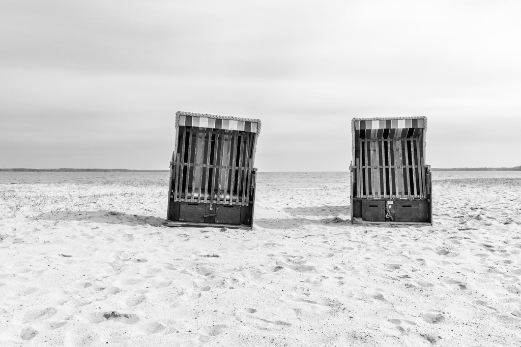 Wandbild: Strandkörbe am Strand in Eckernförde | Dieses Wandbild im Querformat zeigt zwei Strandkörbe am Strand in Eckernförde in Schwarz-Weiß. Der Himmel ist größtenteils bewölkt. - Realisiert mit Pictrs.com