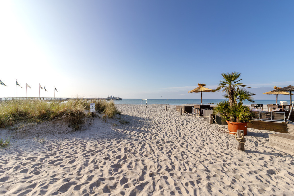 Wandbild: Strandmoment in Grömitz – Zwischen Palmen, Sand und Meer | Dieses Wandbild zeigt eine stilvolle Strandszene in Grömitz an einem sonnigen Tag. Der helle Sand erstreckt sich bis zum ruhigen Meer, das in klarem Blau bis zum Horizont reicht. Links im Bild verläuft die Seebrücke mit wehenden Fahnen. Rechts im Bild befindet sich ein Lounge-Bereich mit Holzmöbeln und strohgedeckten Sonnenschirmen, eingerahmt von dekorativen Palmen in großen Pflanzkübeln. Die Szene wirkt tropisch, geordnet und zugleich entspannt – ein Ort für Ruhe, Genuss und Küstenleben. Der Himmel ist klar, das Licht fällt weich über die gesamte Komposition und schafft eine warme, einladende Atmosphäre. Dieses Motiv eignet sich ideal als Wandbild für maritime Wohnkonzepte – ob als Leinwandbild, Acrylglasbild, Alu-Dibond FineArt Print oder als Akustikbild. Ein stilvoller Akzent für Wohnzimmer, Büro oder Ferienwohnung. - Realisiert mit Pictrs.com