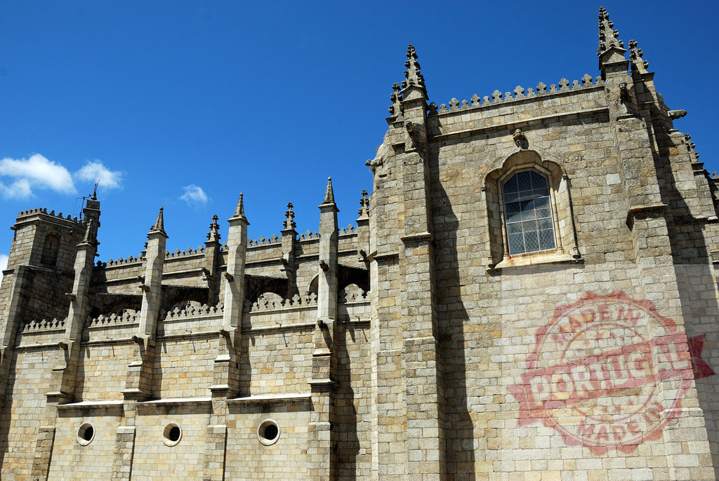 Porto Cathedral flying buttresses and gargoyles against blue sky. Region Norte - Portugal | Porto Cathedral displaying gothic architecture details, flying buttresses, and a stone wall under a clear sky - Realized with Pictrs.com