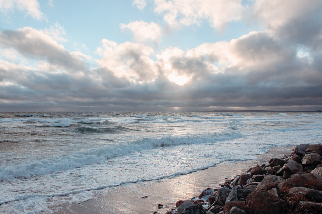Wandbild: Wellen, Wolken und Sonnenschein in dezenten Farben | Dieses Wandbild im Querformat zeigt Wellen und Gischt am Strand in überwiegend hellen Farben. Sowohl am Strand als auch im Meer ist viel weißer Schaum von der Gischt. Dadurch hat das Wandbild viele helle Bestandteile. Am Strand wird das Beige des Sandstrands von der Sonne angestrahlt. Der Himmel auf diesem Bild ist auch sehr malerisch. Am hellblauen Himmel sind graue Wolken, die Teilweise vom roten Licht der aufgehenden Sonne angeleuchtet werden. Kaufen Sie sich diese schöne maritime Morgenstimmung auf Leinwand, Aluminium-Platte oder Acrylglas. Ideal fürs Wohnzimmer, Schlafzimmer, Küche, den Arbeitsplatz oder die Ferienwohnung. Die Wandbilder werden individuell für Sie in vielen Abmessungen produziert. Daher passen die Ostseekult Wandbilder immer perfekt an Ihre Wände. - Realisiert mit Pictrs.com