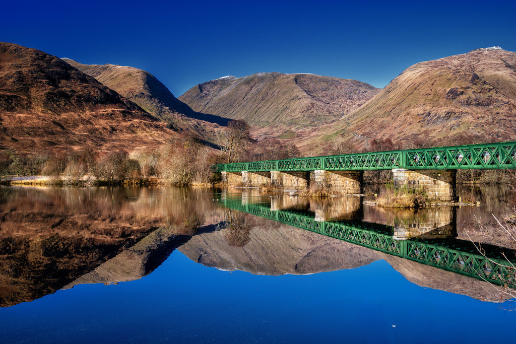Dalmally: Grüne Brücke, Bergreflexion. | Grüne Eisenbahnbrücke über den Fluss Orchy, Dalmally, Schottland, mit Wasserreflexion.A kenspeckle green railway brig gaes ower the lown River Orchy in Dalmally, Scotland. The surroondin Scots Highlands are brichtly mirrored i the water, while the brig maks a naitural sae-side balance. The scene is ruled bi a deep blue lift an the yirdy colours o the winterly hills.Eine markante grüne Eisenbahnbrücke überquert den ruhigen Fluss Orchy in Dalmally, Schottland. Die umliegenden schottischen Highlands spiegeln sich klar im Wasser wider, während die Brücke eine symmetrische Komposition bildet. Die Szene wird von einem tiefblauen Himmel und den erdigen Farben der winterlichen Berglandschaft dominiert. - Realisiert mit Pictrs.com