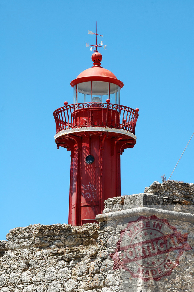 Red lighthouse standing tall on historical stone wall | Red lighthouse tower with a weather vane against a clear blue sky - Realized with Pictrs.com