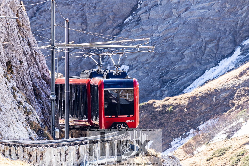 Pilatusbahn am Fels | <p><strong>Technik. Fels. Innovation.</strong></p><p>Die steilste Zahnradbahn der Welt schraubt sich unaufhaltsam durch die massive Felslandschaft am Pilatus. Das leuchtende Rot der modernen Kabine bildet einen faszinierenden Kontrast zum rauen, grauen Stein des Bergmassivs.</p><p>Wähle unter "Produktauswahl" dein Wunschformat: Vom klassischen Wandbild über Puzzle & Tassen bis zum digitalen Download (z.B. als Handy-Hintergrund).</p> - Realisiert mit Pictrs.com