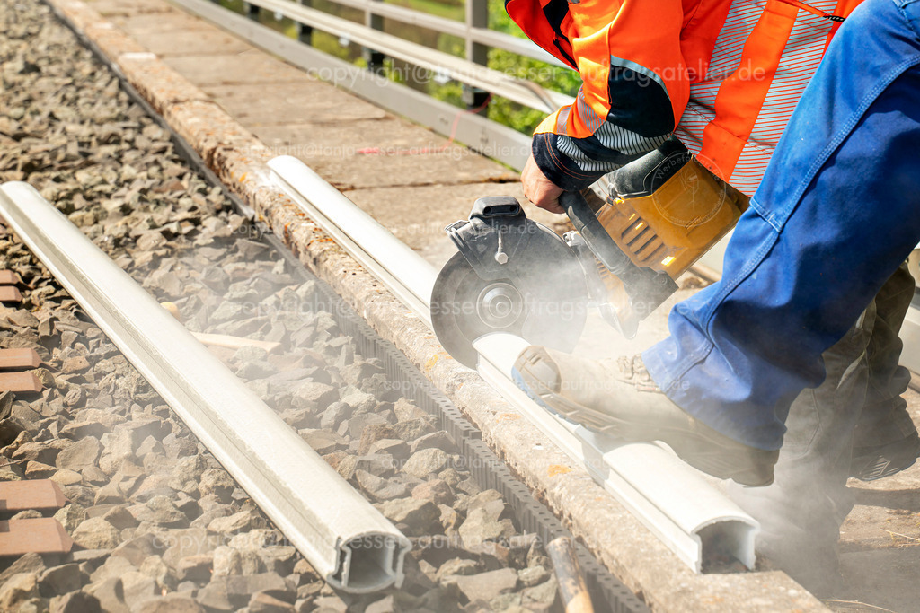Nahaufnahme von zwei Arbeitern  | Ein Arbeiter in Arbeitskleidung und Warnweste trennt mit einer Handkreissäge ein Stück eines Kabelkanals aus Kunststoff ab. Ein Kollege mit Arbeitskleidung hält das Werkstück mit dem Fuß fest. Ein Kabelkanal wird an einem Brückengeländer installiert hoch oben in der Höhe.