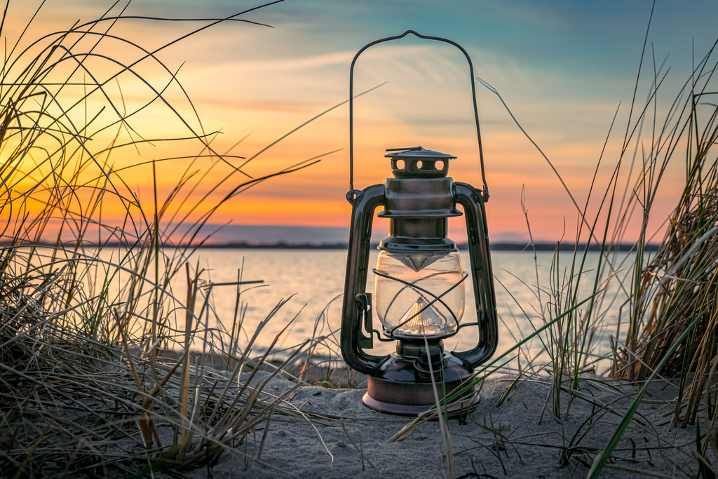 Abendstimmung am Strand | Auf dem Foto ist eine rustikale, antik aussehende Öllampe zu sehen, die zwischen den sanften Hügeln des Dünengrases aufgestellt ist. Das warme, goldene Licht der untergehenden Sonne färbt den Himmel in ein atemberaubendes Spektrum von Orange und Rot, während es sanft auf die Lampe fällt und ihre metallischen Akzente zum Glänzen bringt.