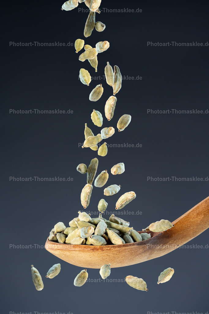 Grüner Kardamom – Aromatische Exotik für feine Genussmomente | as Foto zeigt ganze grüne Kardamomkapseln, die in einem feinen Strahl aus der Bildmitte herabfallen und sich in einem naturbelassenen Holzlöffel sammeln. Jede längliche Kapsel ist gestochen scharf abgebildet und zeigt die charakteristischen Längsrillen sowie die leicht variierenden Grüntöne. Der ruhige, mitteldunkle Grauton des Hintergrunds hebt die organischen Formen und die warme Holzmaserung des Löffels plastisch hervor.TechnikHigh-Speed-Makrofotografie mit sehr kurzer Verschlusszeit: Die fallenden Kapseln sind präzise eingefroren, selbst kleinste Partikel bleiben sichtbar.Geringe Tiefenschärfe: Nur die Kardamomkapseln im Strahl und im Löffel sind scharf; Vorder- und Hintergrund laufen ins sanfte Bokeh über.Weiches, diffuses Licht: Betont Textur und Form der Kapseln, ohne harte Schatten oder ausgebrannte Highlights.FarbenZartes Grün bis Gelbgrün der KardamomkapselnWarme Honig- und Walnussnuancen im HolzlöffelNeutrales Mittelgrau im Hintergrund für klaren, modernen KontrastEmpfohlene Orte zum AufhängenKüche oder Esszimmer – als elegantes Gewürzmotiv, das Inspiration für kreatives Kochen weckt.Asia­restaurant oder Café – ideal über der Bar oder Zubereitungszone, um fernöstliche Aromen zu unterstreichen.Gewürzfachgeschäft oder Feinkostladen – als visueller Eyecatcher, der die Hochwertigkeit und Exotik des Sortiments betont. - Realisiert mit Pictrs.com