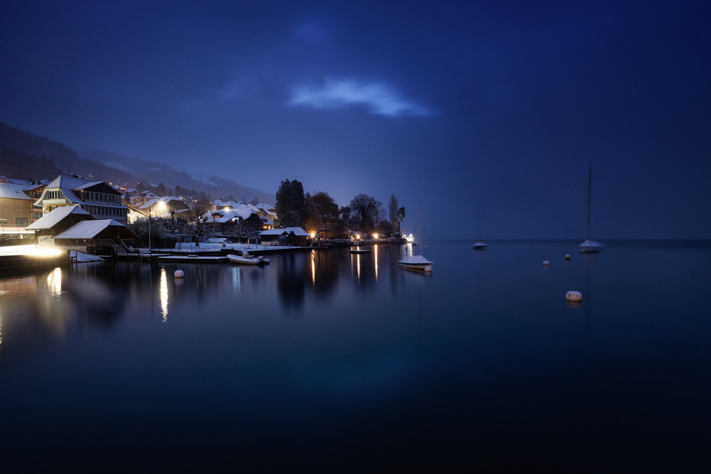 Winterliche Dämmerung in Hilterfingen | Eine winterliche Szene des Dorfes Hilterfingen am Thunersee zeigt beleuchtete Häuser und Bootshäuser mit schneebedeckten Dächern. Die ruhige Wasseroberfläche spiegelt die warmen Lichter wider, während im Hintergrund die dunklen Umrisse der Berge zu erkennen sind. Die Langzeitbelichtung erzeugt eine glatte Wasseroberfläche und betont die atmosphärische Stimmung der blauen Stunde. - Realisiert mit Pictrs.com