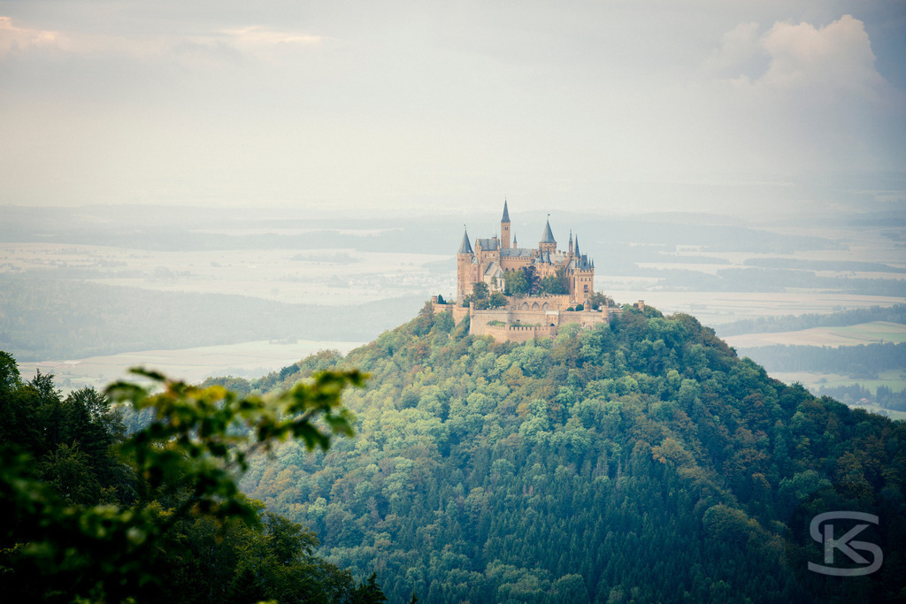 Blick auf die Burg Hohenzollern vom Zeller Horn | Ein malerischer Panoramablick auf die berühmte Burg Hohenzollern auf der Schwäbischen Alb in Deutschland. Das Bild zeigt das dichte Waldgebiet im Vordergrund, das sich über die Hügel erstreckt, und die imposante mittelalterliche Burg auf der Spitze eines markanten Kegelbergs in der Ferne. Die Szene fängt die historische Atmosphäre und die schöne Landschaft der Region ein. - Realisiert mit Pictrs.com