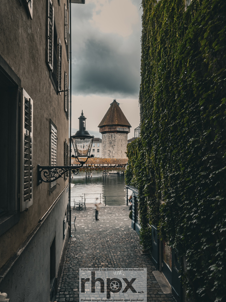 Kapellbrücke Luzern – Blick durch die Altstadtgasse / Chapel Bridge Lucerne – View through the Old Town Alley | Aufnahme aus der Luzerner Altstadt mit Sicht auf die Kapellbrücke und den Wasserturm. Das enge Gassenbild mit den begrünten Hausfassaden zeigt den historischen Charakter der Stadt. *************** A photograph from Lucerne’s old town with a view of the Chapel Bridge and the Water Tower. The narrow alley with its green façades reflects the historic charm and timeless atmosphere of the city. - Realisiert mit Pictrs.com
